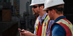 Two contractors on a jobsite wearing safety gear review data on a smartphone.